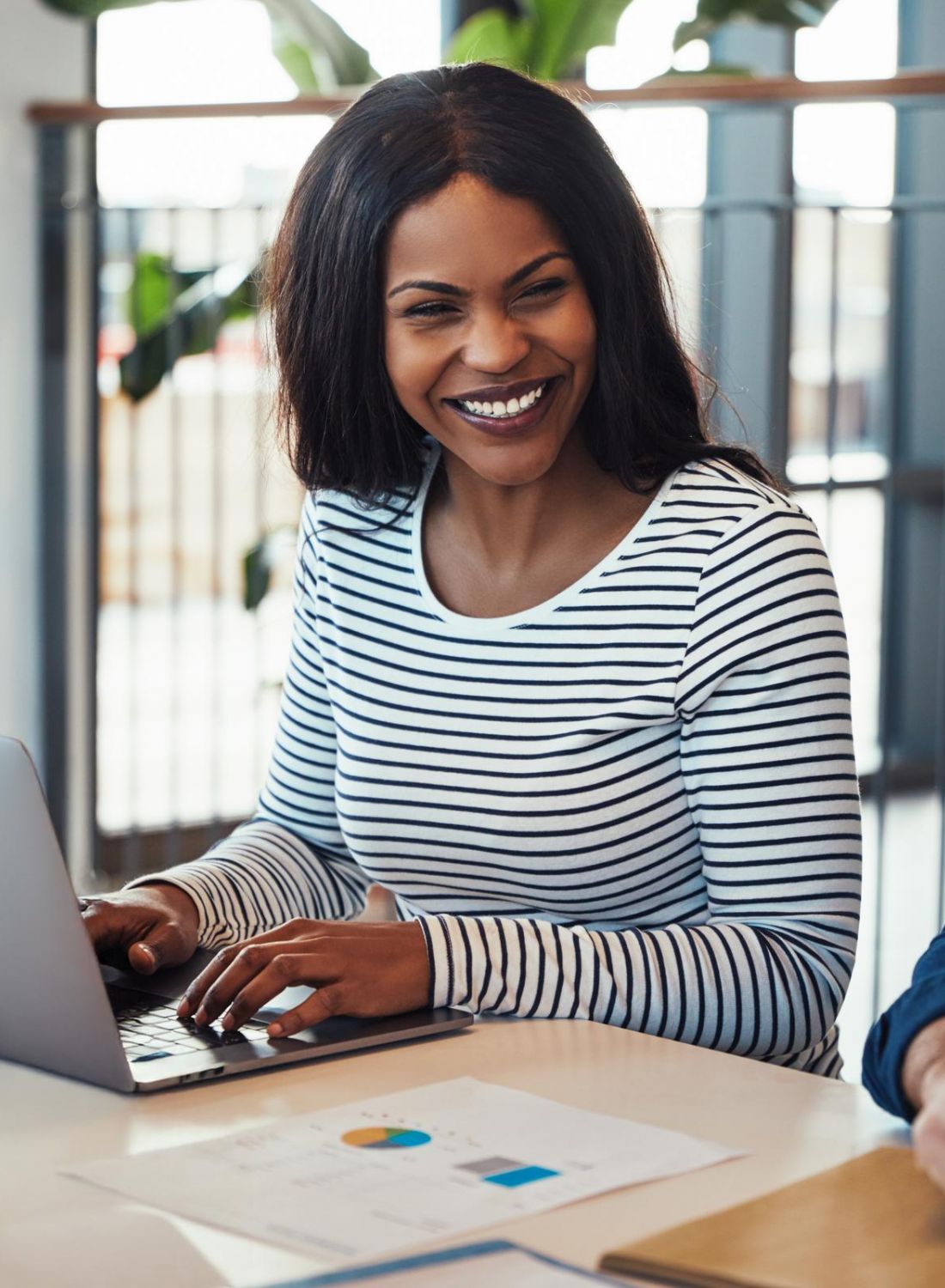 Smiling woman using a laptop at a desk with papers, sitting next to a coworker in an office setting.