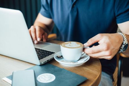 Person using a laptop at a café table, holding a cup of coffee with latte art.