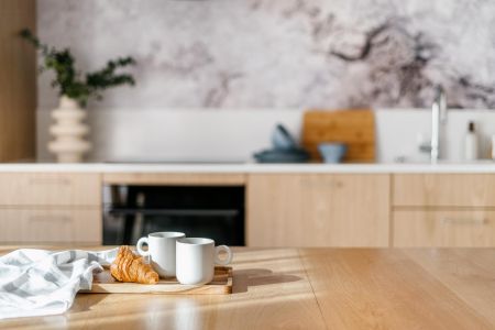 Two white mugs and a croissant on a wooden tray in a modern, sunlit kitchen with light wood cabinets.