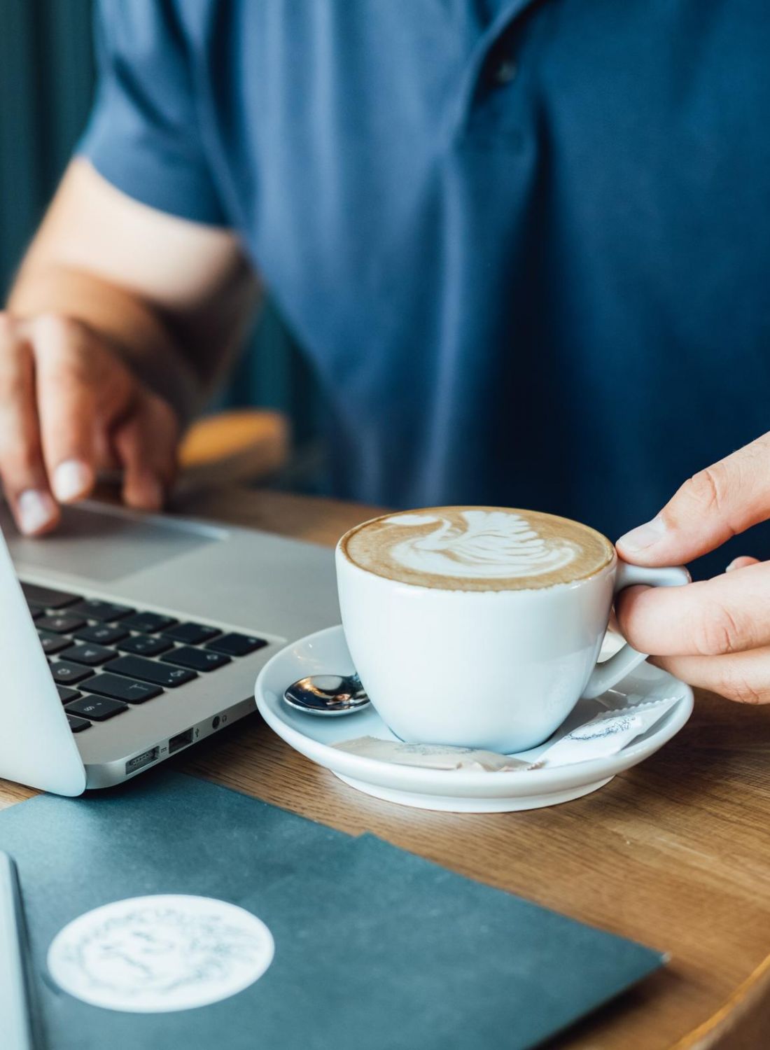 Person using a laptop at a cafe table, holding a cup of coffee with latte art.