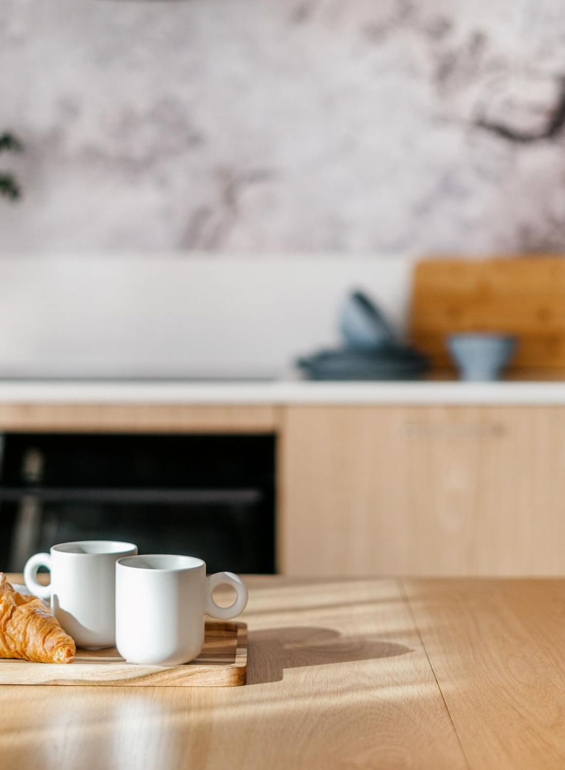 Two white mugs and a croissant on a wooden tray in a modern, sunlit kitchen with light wood cabinets.