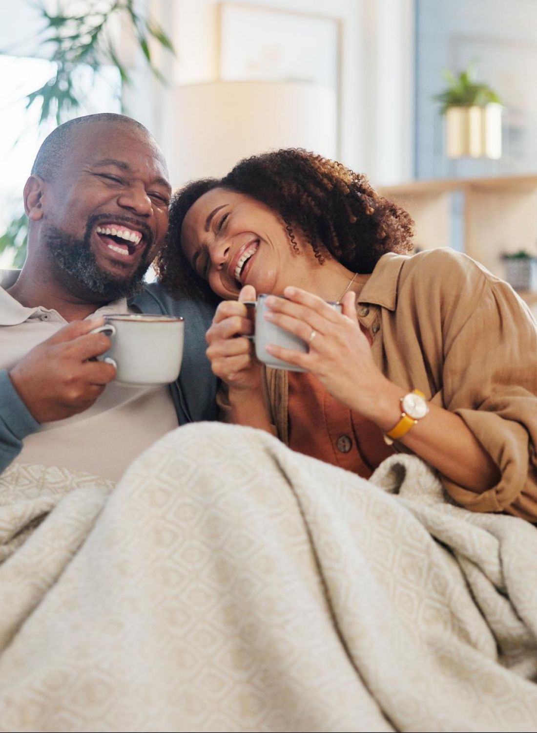 A smiling couple sits on a couch under a blanket, enjoying hot drinks and laughing together.