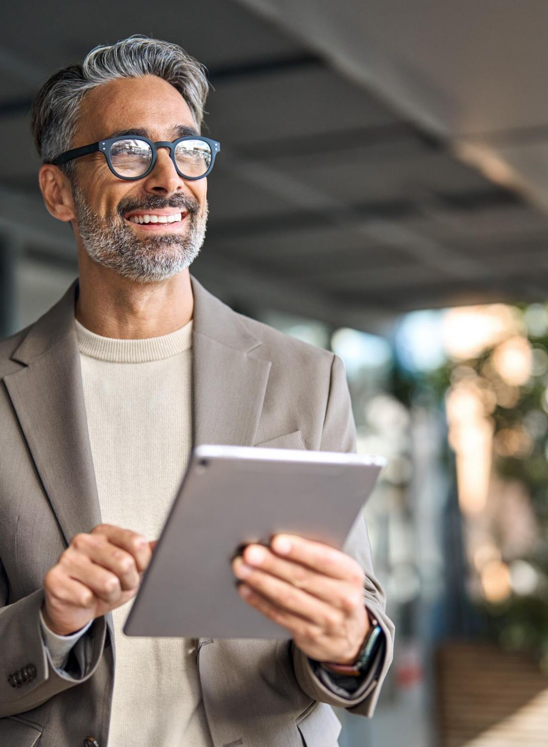 Smiling man in glasses and a blazer using a tablet outdoors in a modern, sunlit setting.