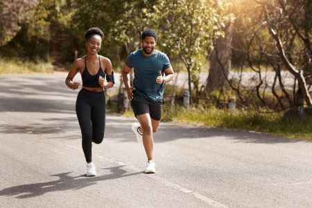 Two people jog together on a sunny, tree-lined road, smiling and enjoying their outdoor run.