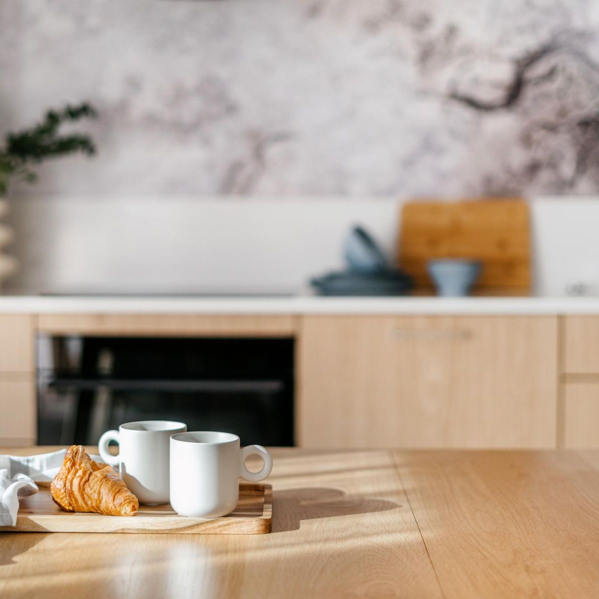 Two white mugs and a croissant on a wooden tray in a modern, sunlit kitchen with light wood cabinets.
