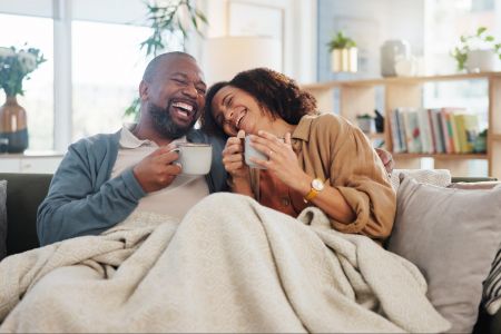 A smiling couple sits on a couch under a blanket, enjoying hot drinks and laughing together.