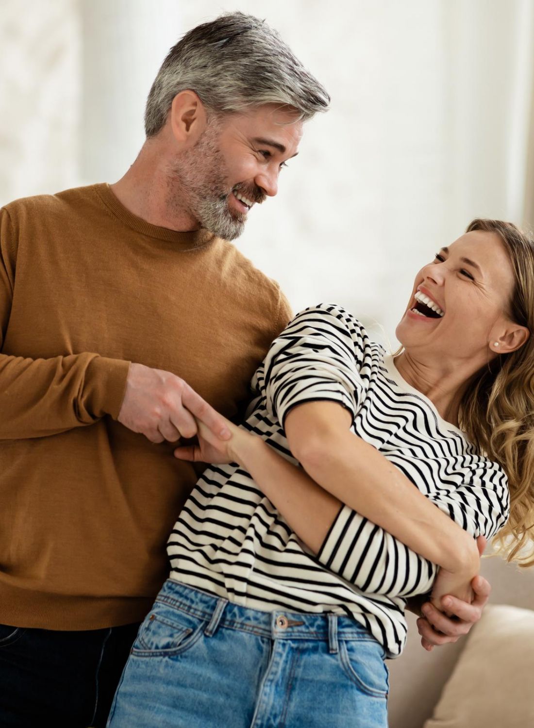 A smiling couple stands together indoors, playfully holding each other and laughing.