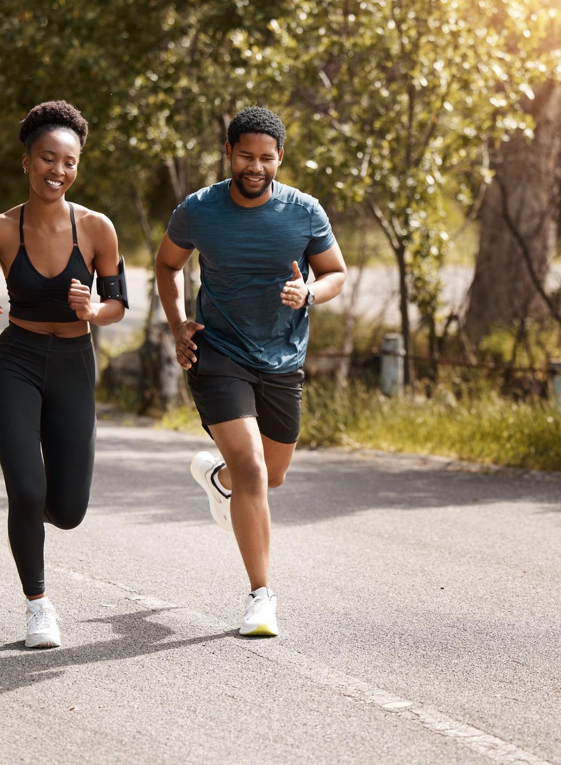 Two people jog together on a sunny, tree-lined road, smiling and enjoying their outdoor run.