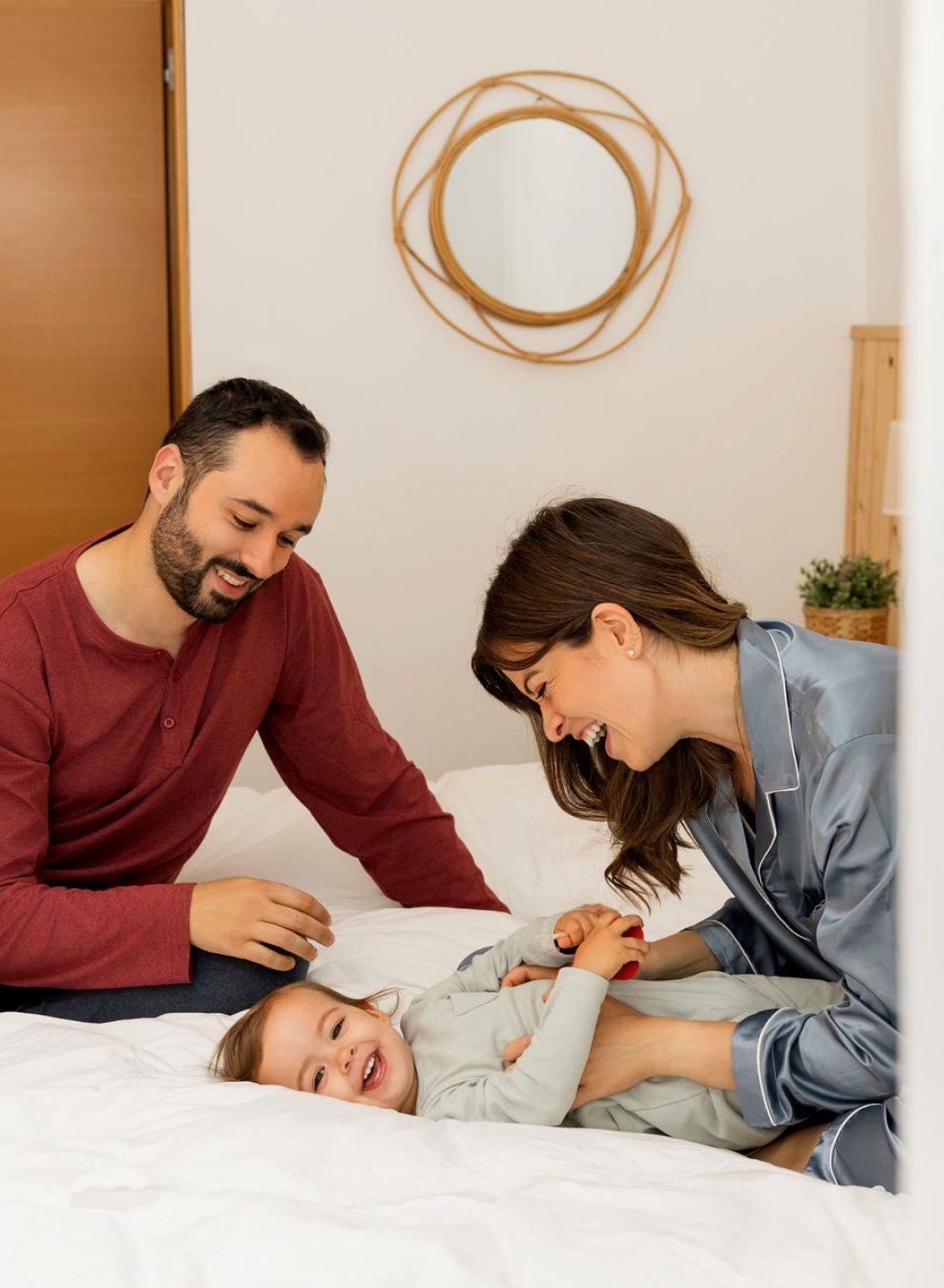 Smiling family of three playing and laughing together on a bed in a bright, cozy bedroom.