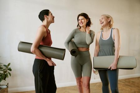 Three women laughing and holding yoga mats, standing together in workout clothes indoors.